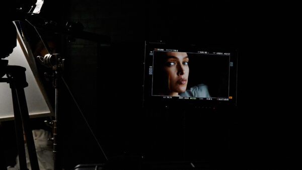 Monitor on a dim film set showing a close-up of a woman's face while lighting and camera gear loom in the background