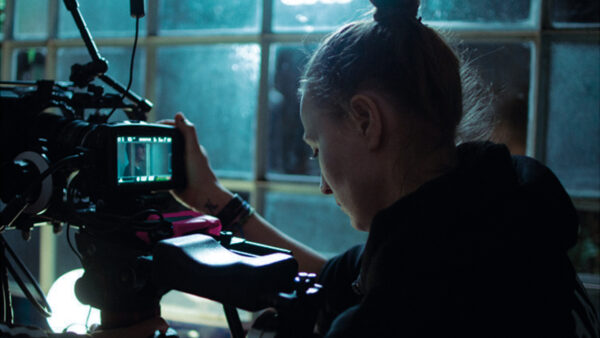 Female videographer adjusting a professional camera on a tripod, indoors by a window.